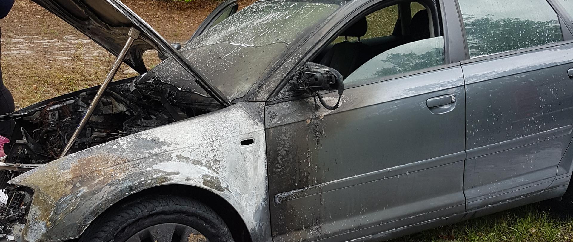 Zdjęcie przedstawia samochód osobowi marki Audi z otwartą maską. Widać ślady po pożarze - wokół znajduje się piana gaśnicza. Przed samochodem znajduje się pachołek ostrzegawczy.