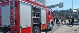 na pierwszym planie samochód pożarniczy dalej grupa strażaków pod koszem drabiny mechanicznej, w tle widać silosy cukrowni