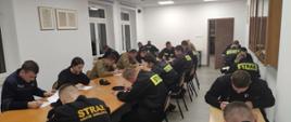 Na zdjęciu strażacy z Ochotniczych Straży Pożarnych w trakcie egzaminu kończącym szkolenie dowódców OSP. Strażacy ubrani w ubrania koszarowe koloru czarnego i piaskowego bez czapek na sali wykładowej siedzą na krzesłach przed owalnym stole i stolikach przy ścianie w Komendzie Powiatowej Państwowej Straży Pożarnej w Gorlicach.