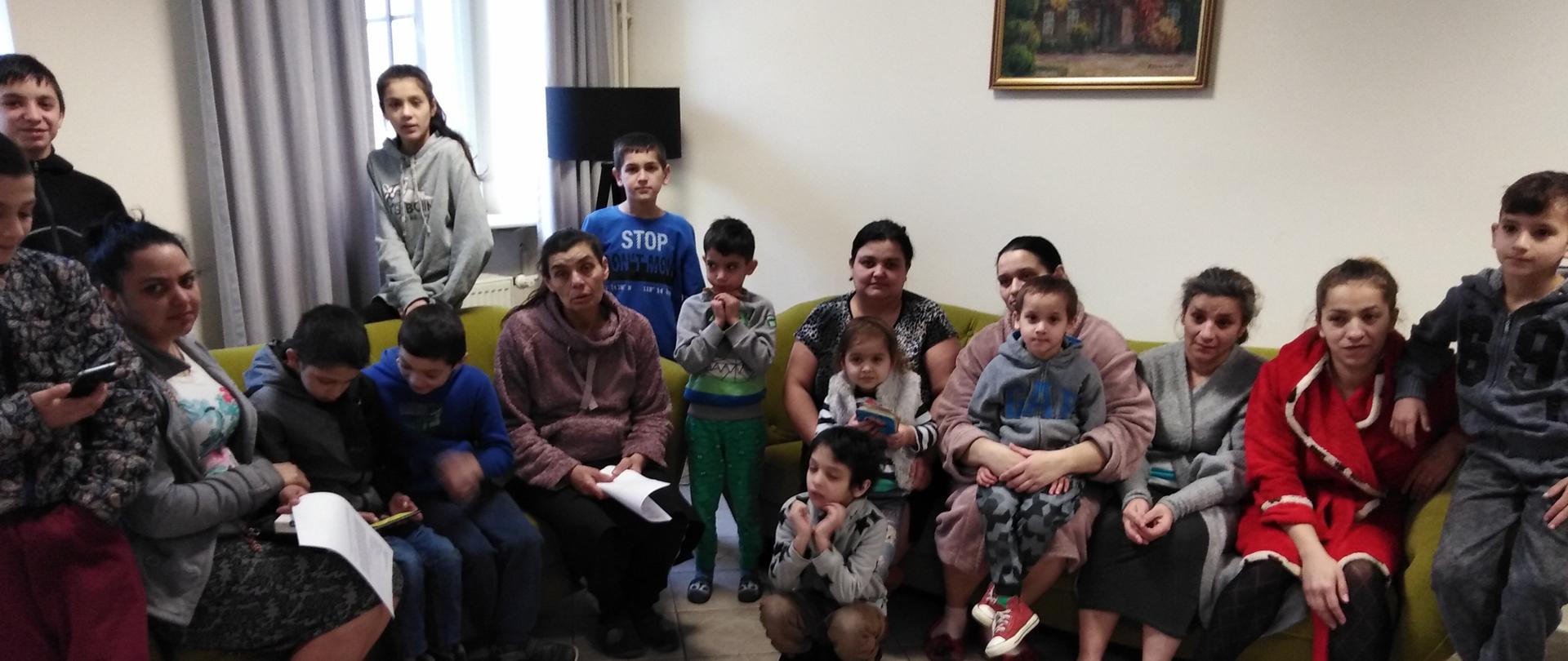 Grupa osób kobiet i dzieci siedzi na dwóch kanapach. Niektóre dzieci siedzą na kolanach matek. 