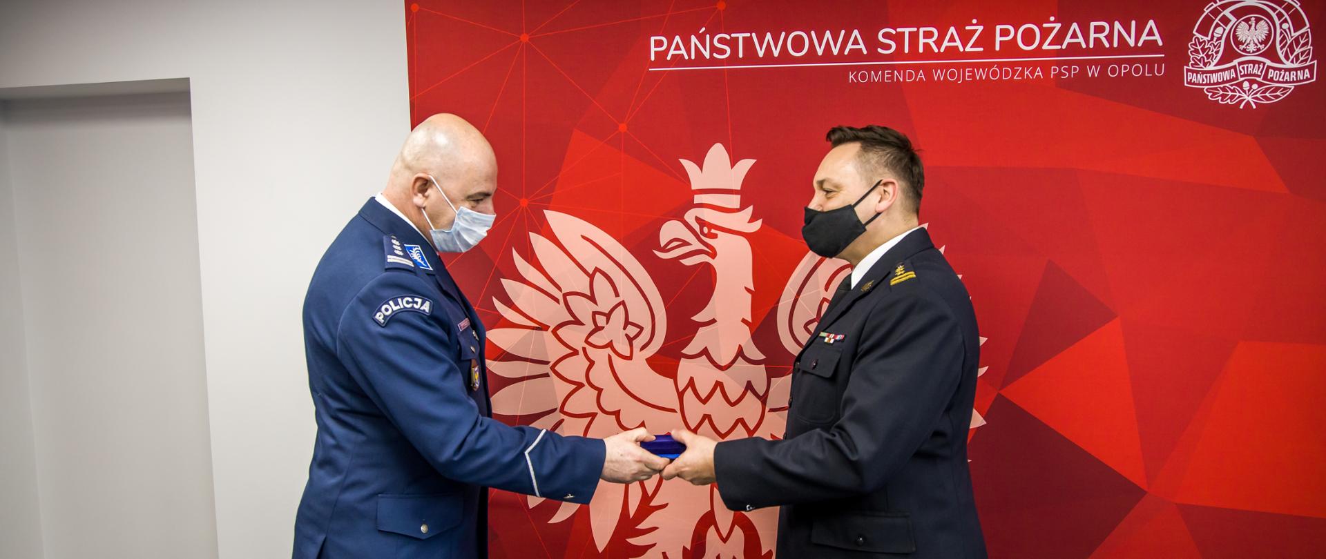 Na zdjęciu insp. Rafał Kochańczyk Komendant Wojewódzki Policji w Opolu wręcza Brązowy Medal za Zasługi dla Policji st. bryg. Krzysztofowi Kędrykowi Opolskiemu Komendantowi Wojewódzkiemu PSP w Opolu. W tle czerwona ścianka z napisem PSP i Komenda Wojewódzka PSP w Opolu wraz z białym orłem.