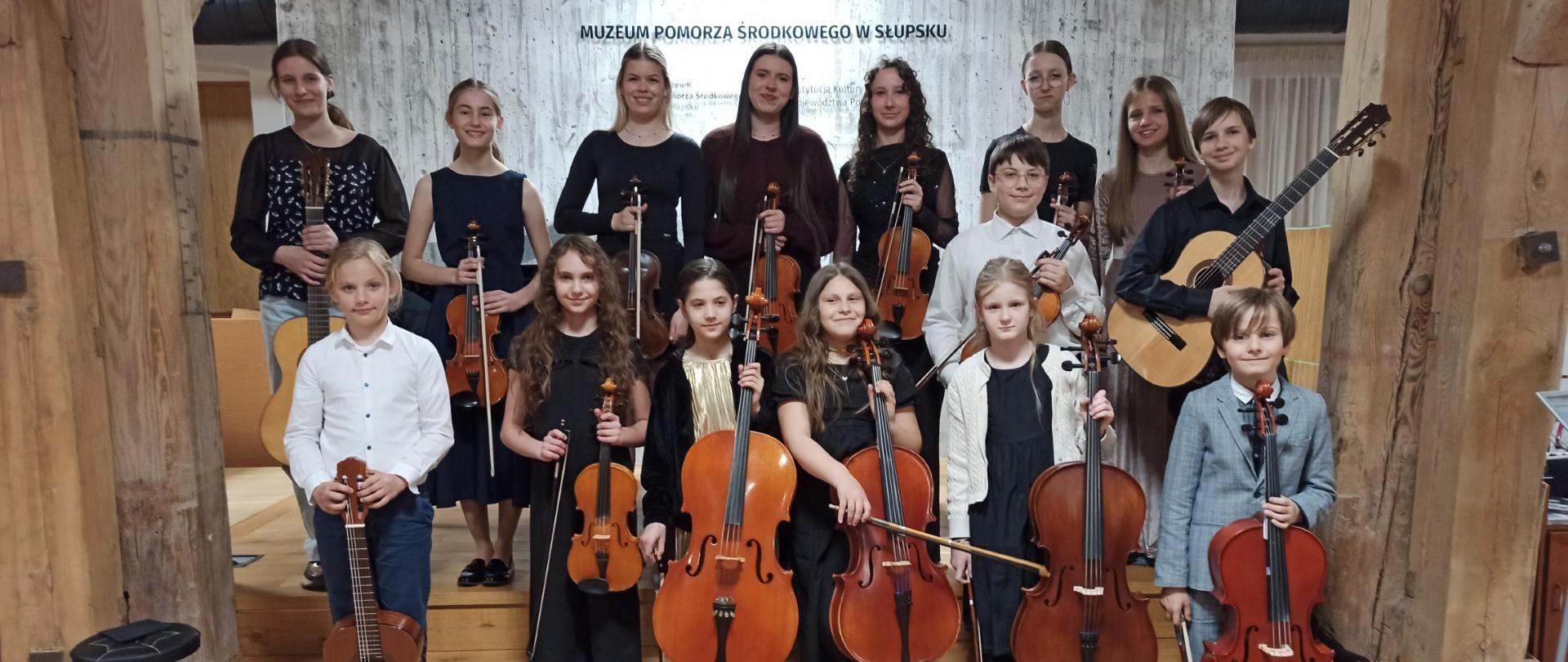 Zdjęcie kolorowe. Grupa kilkunastu osób w różnym wieku, stojąca na drewnianym podwyższeniu w pomieszczeniu o rustykalnym charakterze z grubymi, drewnianymi belkami konstrukcyjnymi. Osoby trzymają instrumenty strunowe: skrzypce, wiolonczele oraz gitary klasyczne. Wszyscy są zwróceni przodem, ubrani w stroje galowe – przeważają kolory czarny, biały i szary. W tle jasna ściana z napisem "MUZEUM POMORZA ŚRODKOWEGO W SŁUPSKU". Podłoga wykonana z jasnych paneli drewnianych, po lewej stronie czarny stołek muzyczny. Oświetlenie jasne, sufitowe.