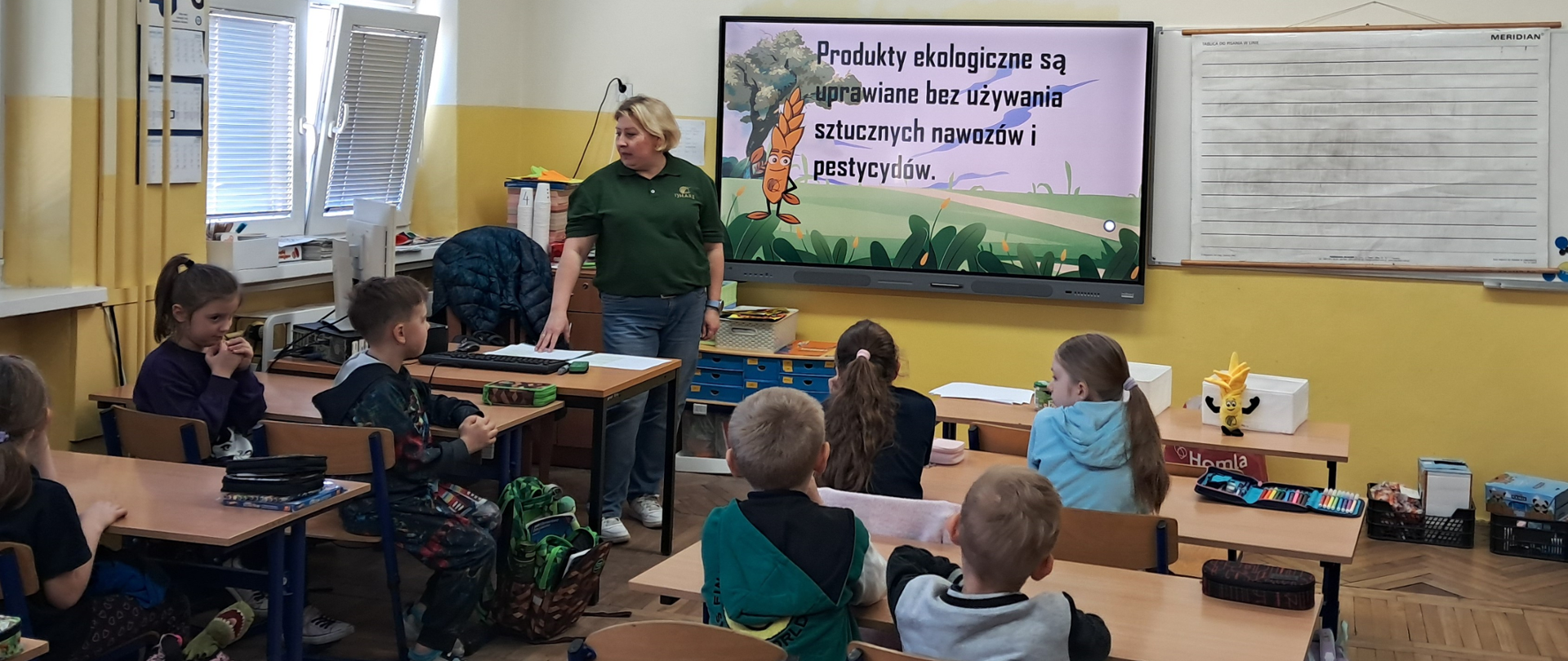 Dzieci siedzą w szkolnych ławkach i uważnie słuchają inspektorki z Inspekcji, która prowadzi lekcję na temat ekologii; w tle widoczna jest prezentacja multimedialna.