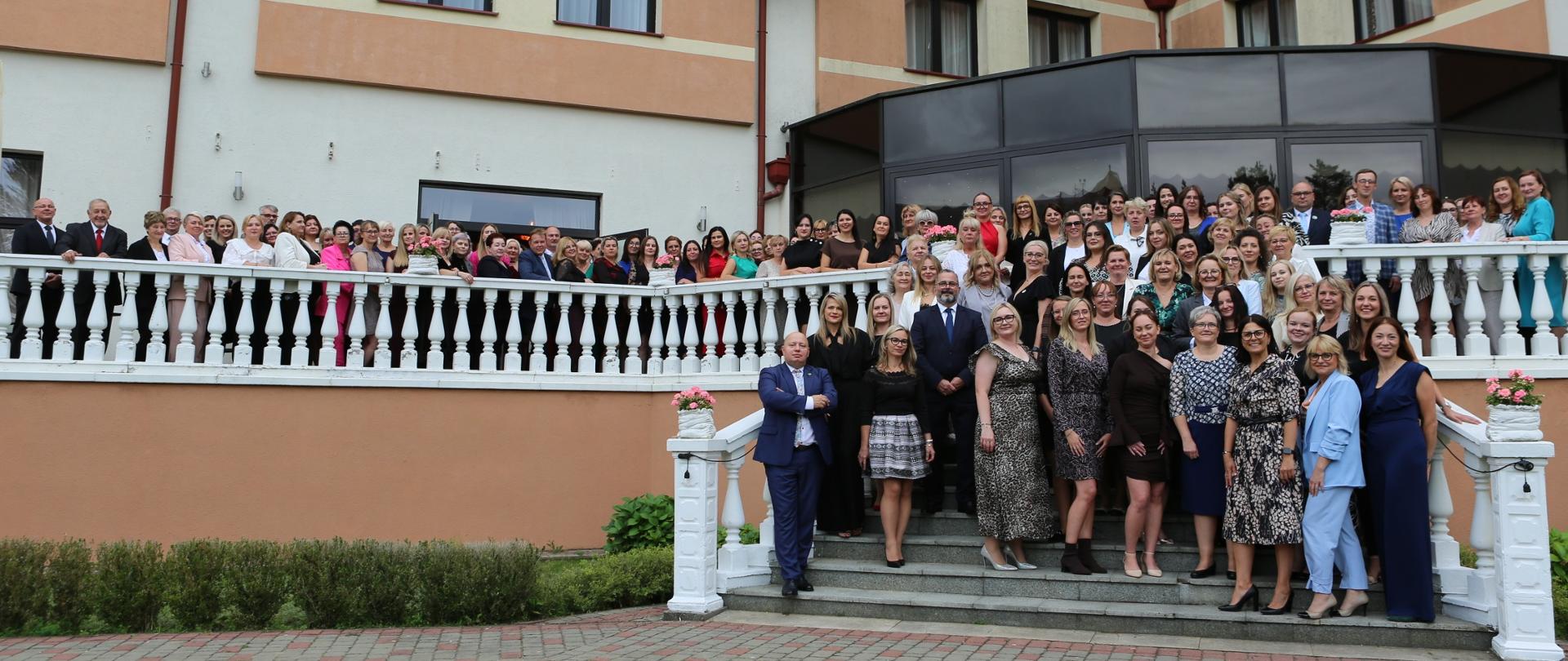 Około setka osób pozuje do zdjęcia na tarasie i schodach budynku. Wszyscy są odświętnie ubrani i uśmiechają się do zdjęcia.