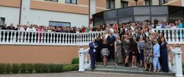 Około setka osób pozuje do zdjęcia na tarasie i schodach budynku. Wszyscy są odświętnie ubrani i uśmiechają się do zdjęcia.