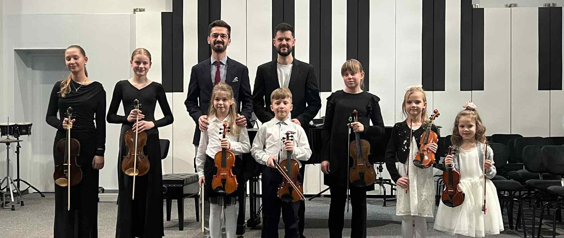 Wszyscy wykonawcy stoją razem z nauczycielem na scenie.