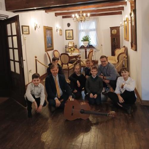 Sześcioro uczniów i nauczyciel siedzą na drewnianej podłodze w salonie u Lutosławskich. Przed nimi położona gitara klasyczna. Za uczniami salonik z meblami z XIX wieku.