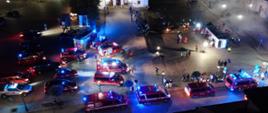 Widok z góry, na płycie rynku samochody pożarnicze z włączoną sygnalizacją świetlną. Sceneria nocna. Działania podczas symulowanego wypadku autobusu.
