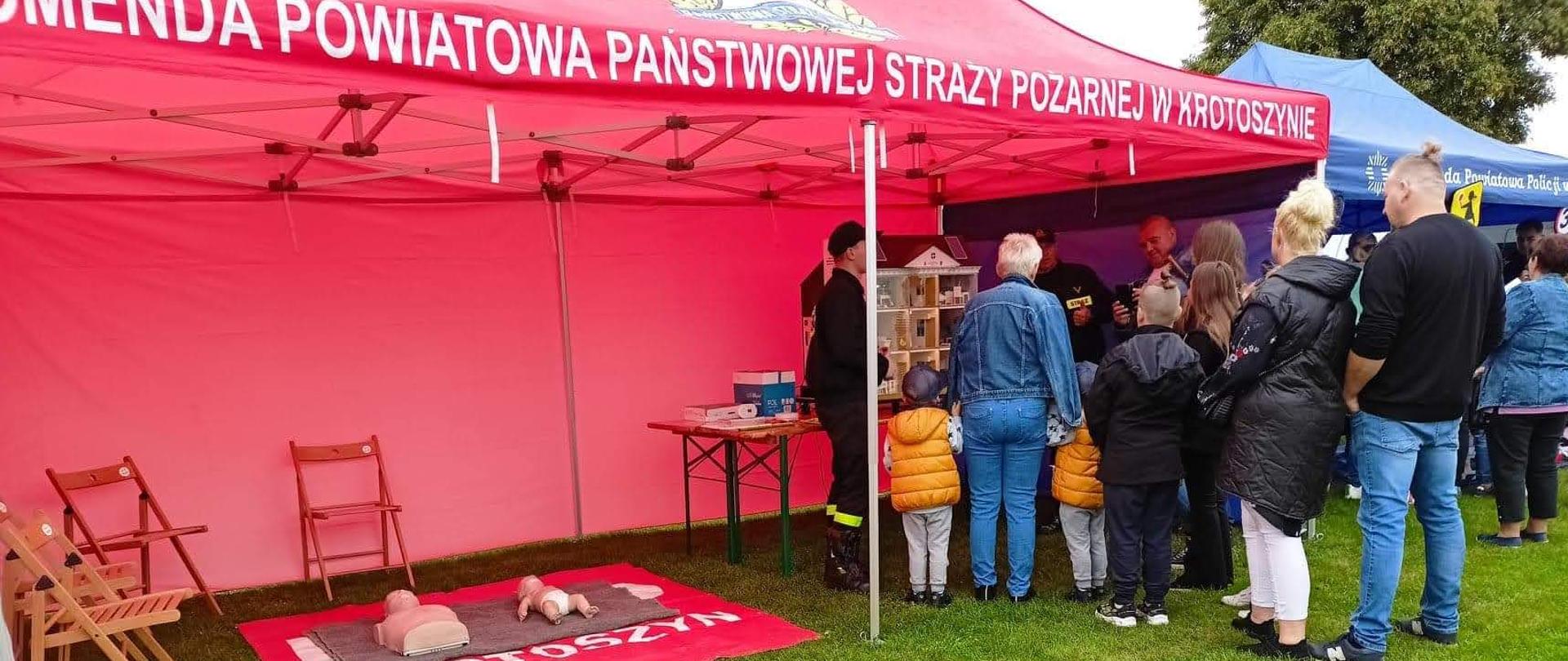 Pod namiotem strażackim, strażacy prezentują mobilny symulator zagrożeń w budynku jednorodzinnym. Uczestnicy dozynek przyglądają się symulacji zagrożeń. Strażacy przedstawiają zagrożenia tłumacząc jak ich uniknąć.