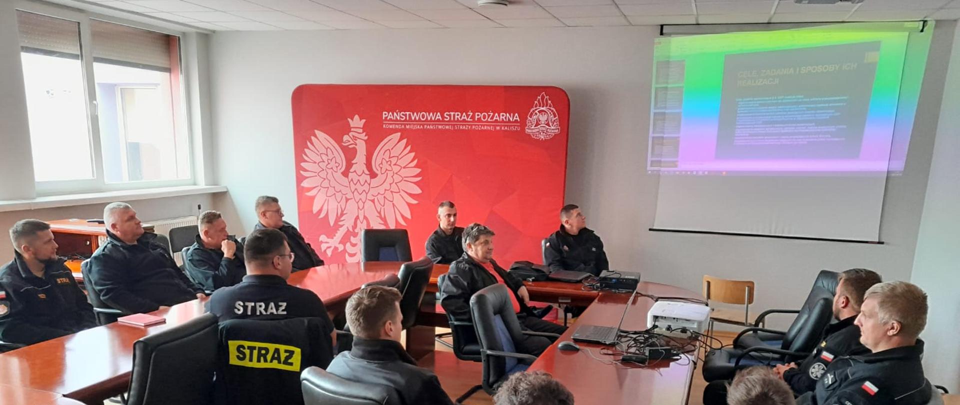 Na zdjęciu widzimy strażaków ochotników ubranych w mundury koszarowe koloru czarnego. Strażacy znajdują się w sali wykładowej, siedzą przy stole, przed nimi biały ekran na którym rzucane są slajdy prezentacji. 