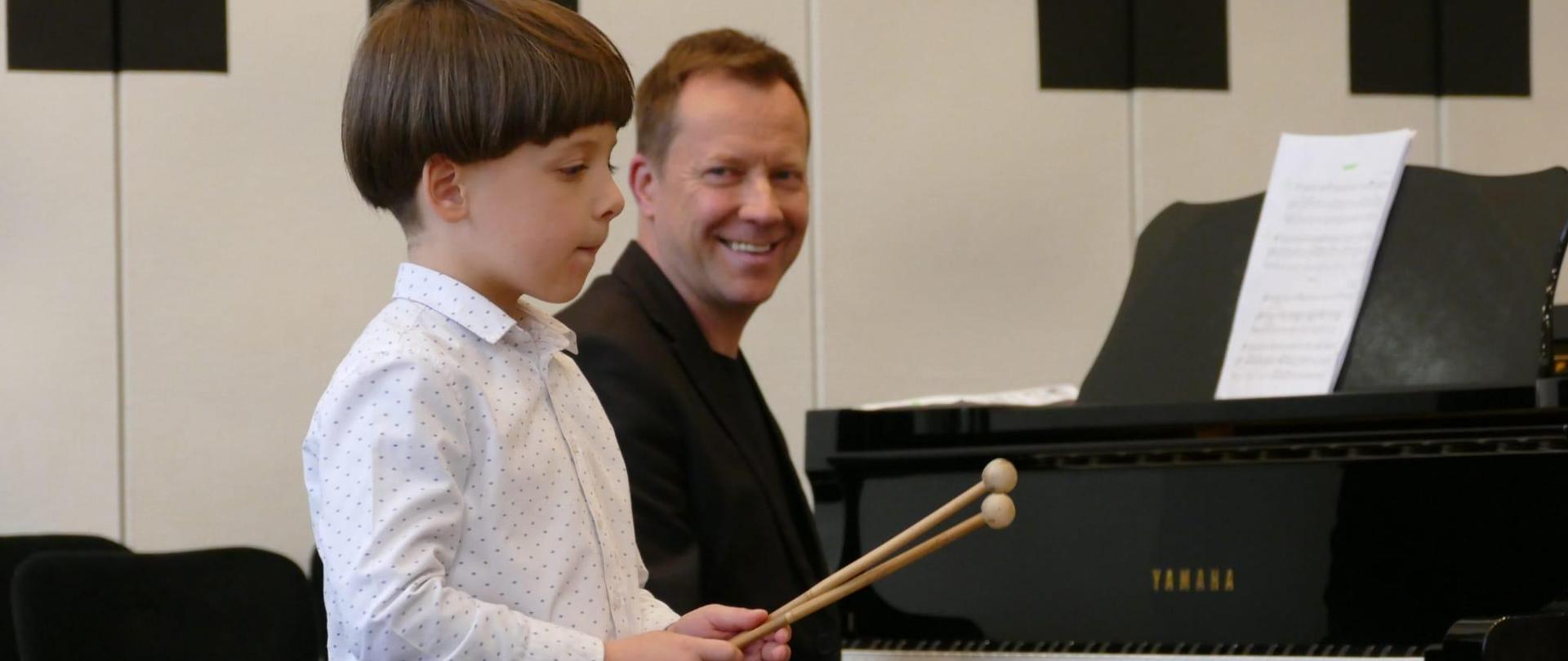 Chłopiec gra na dzwonkach, za nim mężczyzna siedzi przy fortepianie.
