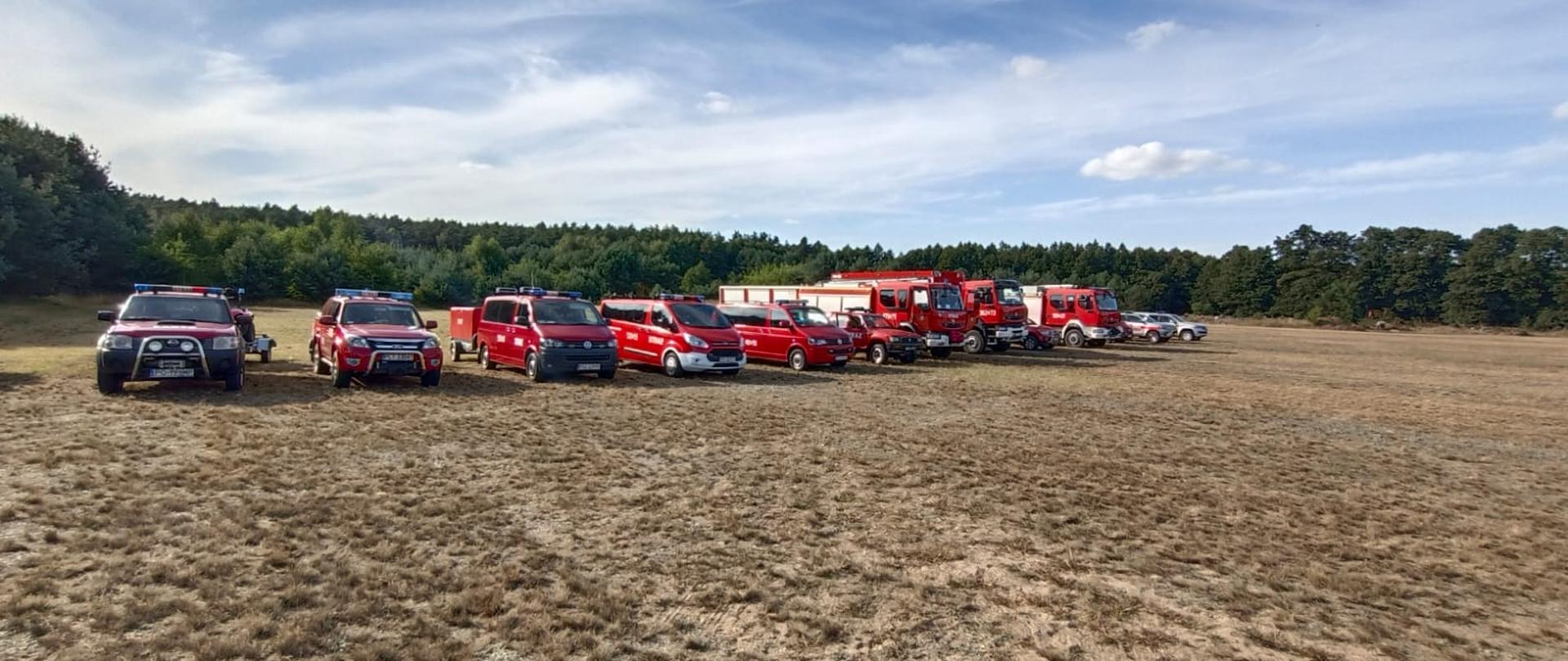 Widać samochody straży na pacu zbiórki, za nimi las