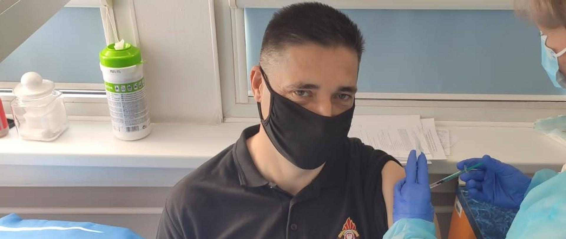 strażak w umundurowaniu dowódczo sztabowym w czarnym podkoszulku przyjmuje szczepionkę