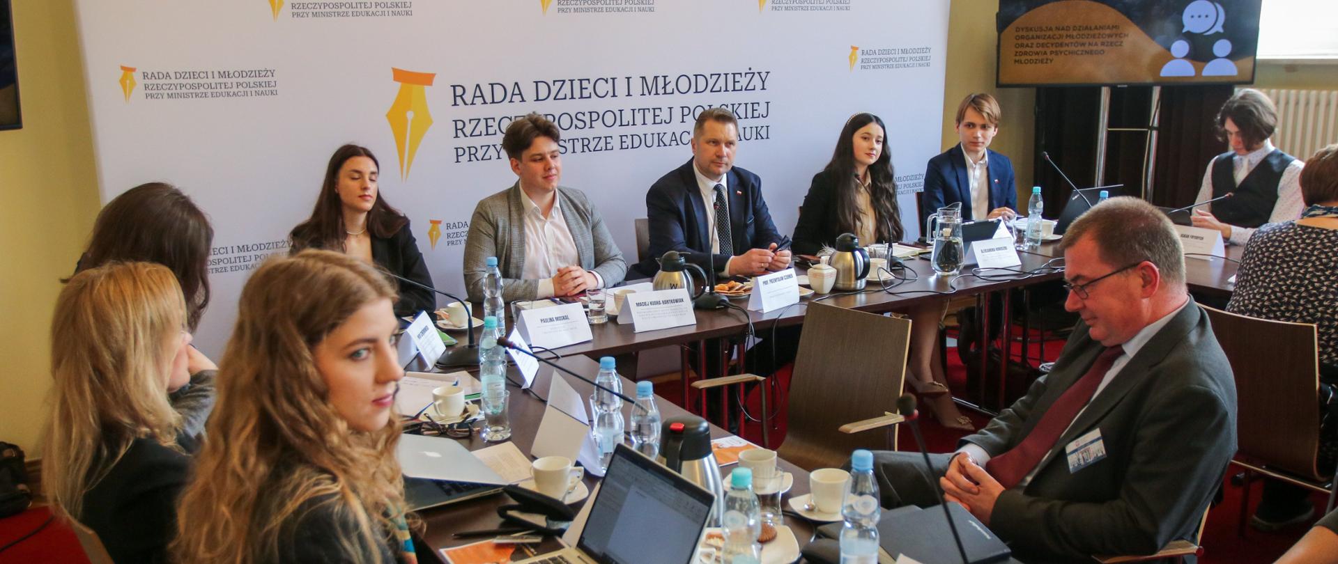 Zdjęcie z góry, w sali przy ustawionych w czworokąt stołach siedzi dużo młodych ludzi, przy jednej ścianie ścianka z napisem Rada dzieci i młodzieży, dalej okno.