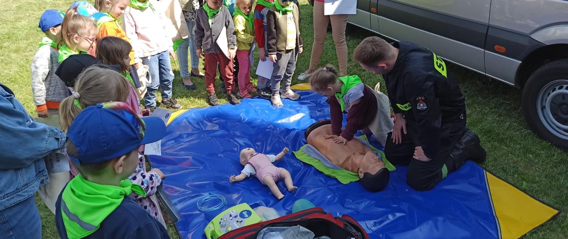Na zdjęciu widać niebieską plandekę rozłożona na trawniku. Na plandece uczestnicy majówki ze strażakiem ćwiczą udzielanie pierwszej pomocy na fantomie. Obok stoi czerwona torba ze sprzętem medycznym. Na trawniku stoją inne osoby, które obserwują osobę ćwiczącą. W tle widać srebrny samochód służby więziennej, wojskowej straży pożarnej w kolorze khaki, budynek oraz boisko. Jest widno.