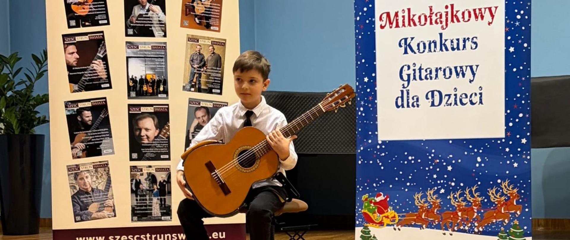 Chłopiec siedzi na scenie z gitarą klasyczną, pozując pomiędzy dwoma dużymi banerami reklamującymi konkurs gitarowy i czasopismo muzyczne. Jest ubrany w białą koszulę z krawatem, a cała scena oświetlona jest ciepłym światłem podkreślającym drewnianą podłogę.