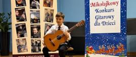 Chłopiec siedzi na scenie z gitarą klasyczną, pozując pomiędzy dwoma dużymi banerami reklamującymi konkurs gitarowy i czasopismo muzyczne. Jest ubrany w białą koszulę z krawatem, a cała scena oświetlona jest ciepłym światłem podkreślającym drewnianą podłogę.
