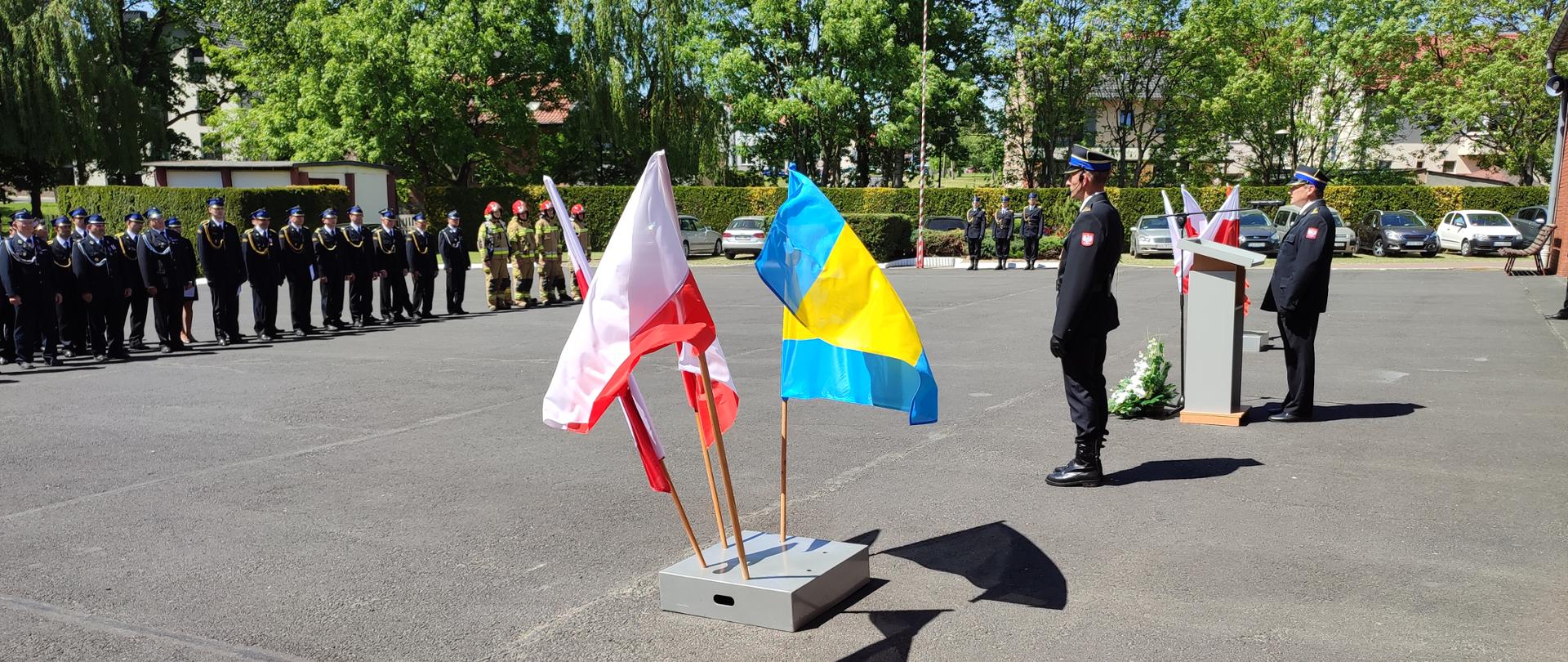 Plac wewnętrzny strażnicy, w tle stoją strażacy w mundurach wyjściowych i specjalnych koloru beżowego. Na środku placu mównica przy której przemawia strażak obok mównicy stoi kolejny strażak oraz powiewają flagi Polski.