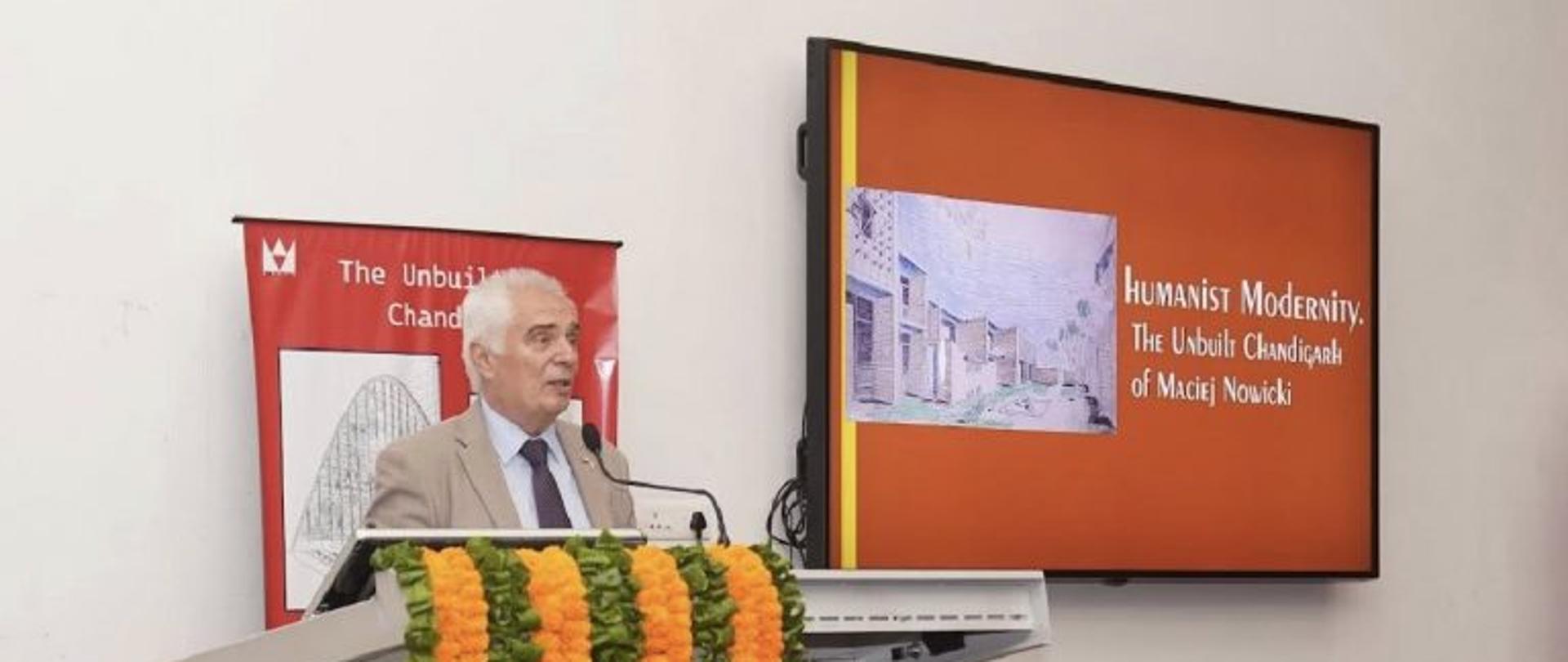 Head of Mission of the Embassy of Poland in New Delhi, Dr Piotr Antoni Świtalski, delivers a speech at a lectern decorated with orange and green flower garlands. A screen beside him displays the title of the exhibition, “Humanist Modernity: The Unbuilt Chandigarh of Maciej Nowicki,” together with a reproduction of an architectural drawing.