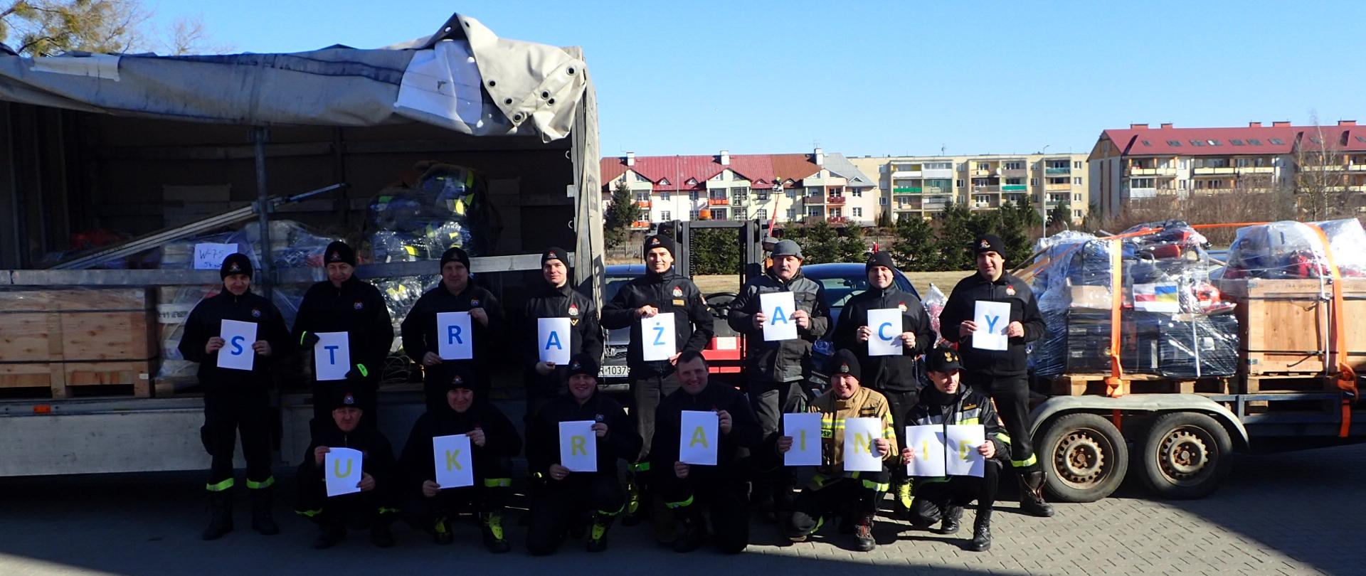 Grupa strażaków stoi z napisem "Strażacy Ukrainie". W tle załadowany na samochody sprzęt pożarniczy. 