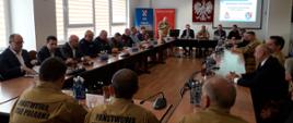 Strażacy oraz zaproszeni goście znajdują się w sali narad. Siedzą za ułożonymi w prostokąt stołami. Przemawia komendant powiatowy Państwowej Straży Pożarnej. Na ekranie wyświetlana jest prezentacja.