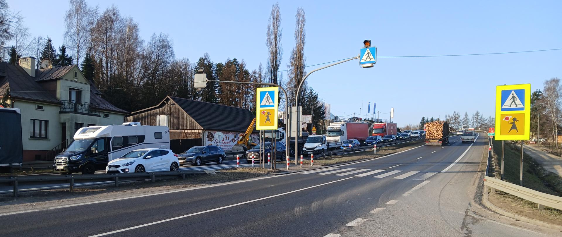 Przejście dla pieszych na dwujezdniowej drodze z wysepką dzielącą, wyposażone w sygnalizację świetlną z ostrzegawczymi sygnałami żółtymi. Widoczne oznakowanie pionowe przejścia oraz logotyp „Aktywny znak”. Na jezdni ruch samochodowy, w tym samochody osobowe i ciężarowe; po lewej stronie budynki mieszkalne i gospodarcze, po prawej pobocze z barierą ochronną.