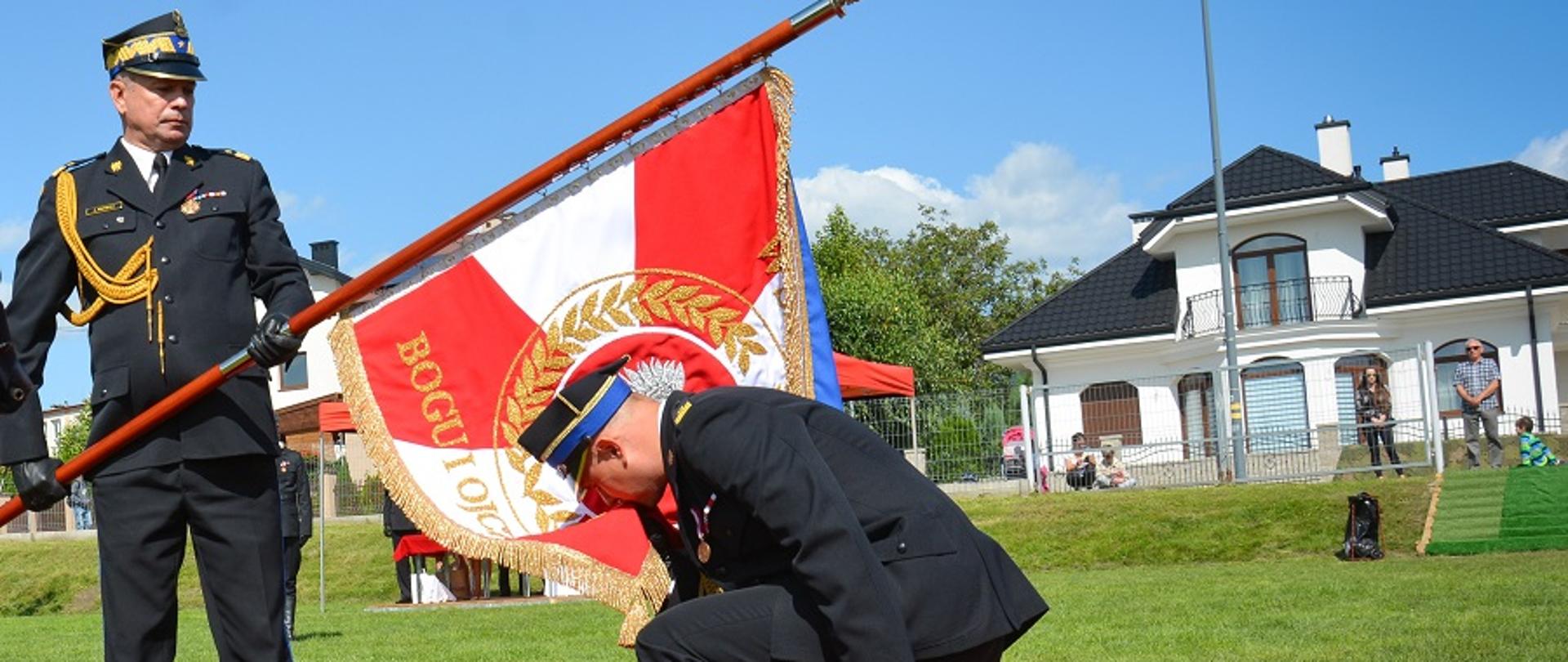 Plac apelowy uroczystości nadania sztandaru komendzie powiatowej Państwowej Straży Pożarnej w Mielcu. Na fotografii widać moment przekazania przez zastępcę komendanta głównego PSP nowo nadanego sztandaru komendzie powiatowej PSP w Mielcu. Na podeście okrytym czerwonym suknem stoi trzymając sztandar zastępca komendanta głównego. Przy sztandarze klęcząc na jedno kolano komendant powiatowy PSP w Mielcu całuje róg sztandaru. Strażacy ubrani są w mundury wyjściowe koloru granatowego. Głowy mają nakryte czapkami rogatywkami.
