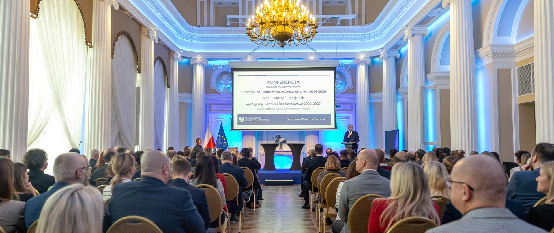 Sala konferencyjna z kolumnami i żyrandolem. Prelegent przemawia na scenie, obok flag Polski i UE. Publiczność siedzi na krzesłach, a na ekranie wyświetlany jest temat konferencji.