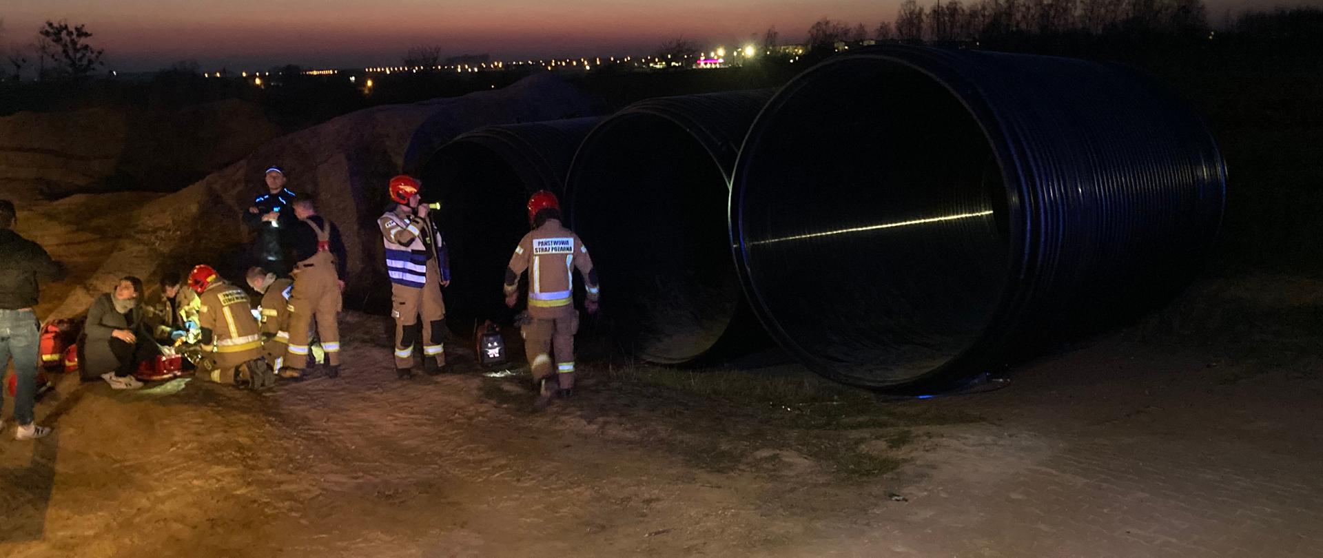Strażacy udzielają KPP dziewczynce. Obok znajdują się bardzo duże rury.