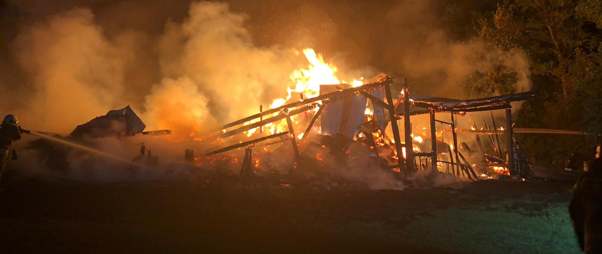 Na środku zdjęcia widzimy powaloną konstrukcje budynku – stodoły w nocy. Konstrukcja budynku cała objęta ogniem, a nad nią unoszące się kłęby dymu oraz iskry. Z pożaru wystają arkusze blachy, które przykrywały budynek. Na środku pożaru widać zmagazynowaną, palącą się słomę. Po lewej stronie widzimy Strażaka ubranego w ubranie specjalnie typu Nomex podającego prąd wody w natarciu na pożar. Po prawej stronie widzimy wysokie drzewa, przy których stoi strażak i podaje drugi prąd wody. Przed palącym się budynkiem widać rozciągniętą linie gaśniczą leżącą na trawie. 