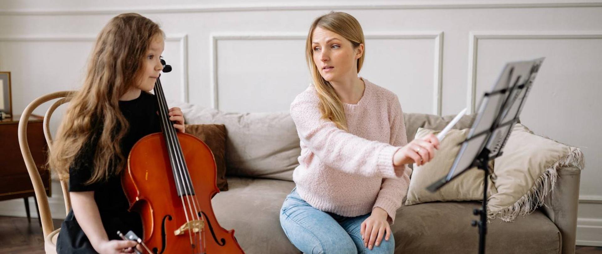 Zjęcia przedstawia po lewej siedzącą dziewczynkę z wiolonczelą, a po prawej nauczycielkę instrumentu tłumaczącą zagadnienia uczennicy.
