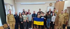 Pamiątkowe zdjęcie uczestników szkolenia. Na pierwszym planie fantomy do nauki i sprzęt do udzielania kwalifikowanej pierwszej pomocy. Jedna z osób trzyma niebiesko-żółtą flagę państwową Ukrainy. 