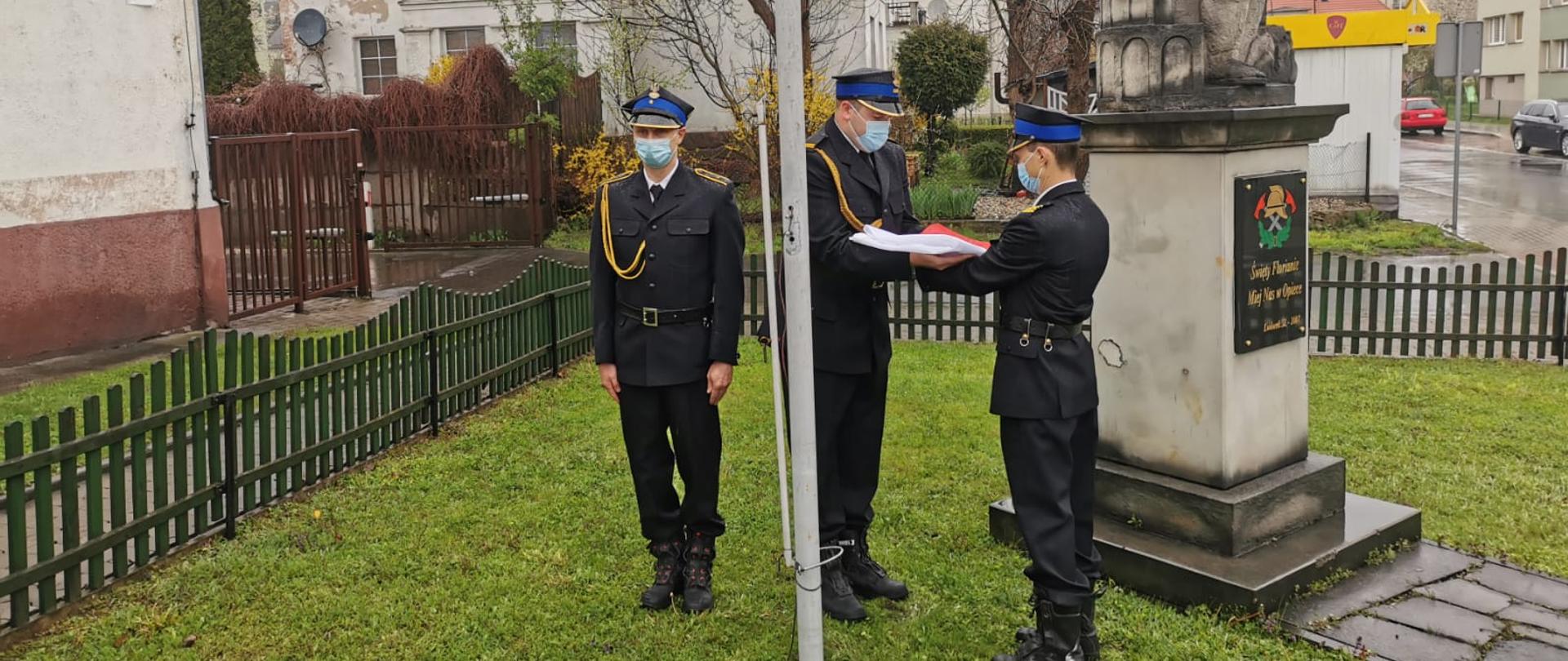 Zdjęcie przedstawia trzech strażaków stojących przy maszcie podczas uroczystej zmiany służby przed podniesieniem flagi państwowej. 