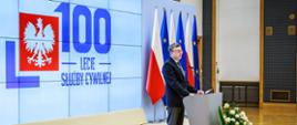 Wystąpienie szefa służby cywilnej Dobrosława Dowiata-Urbańskiego - w tle ekran z logo 100-lecia służby cywilnej oraz flagi Polski i Unii Europejskiej