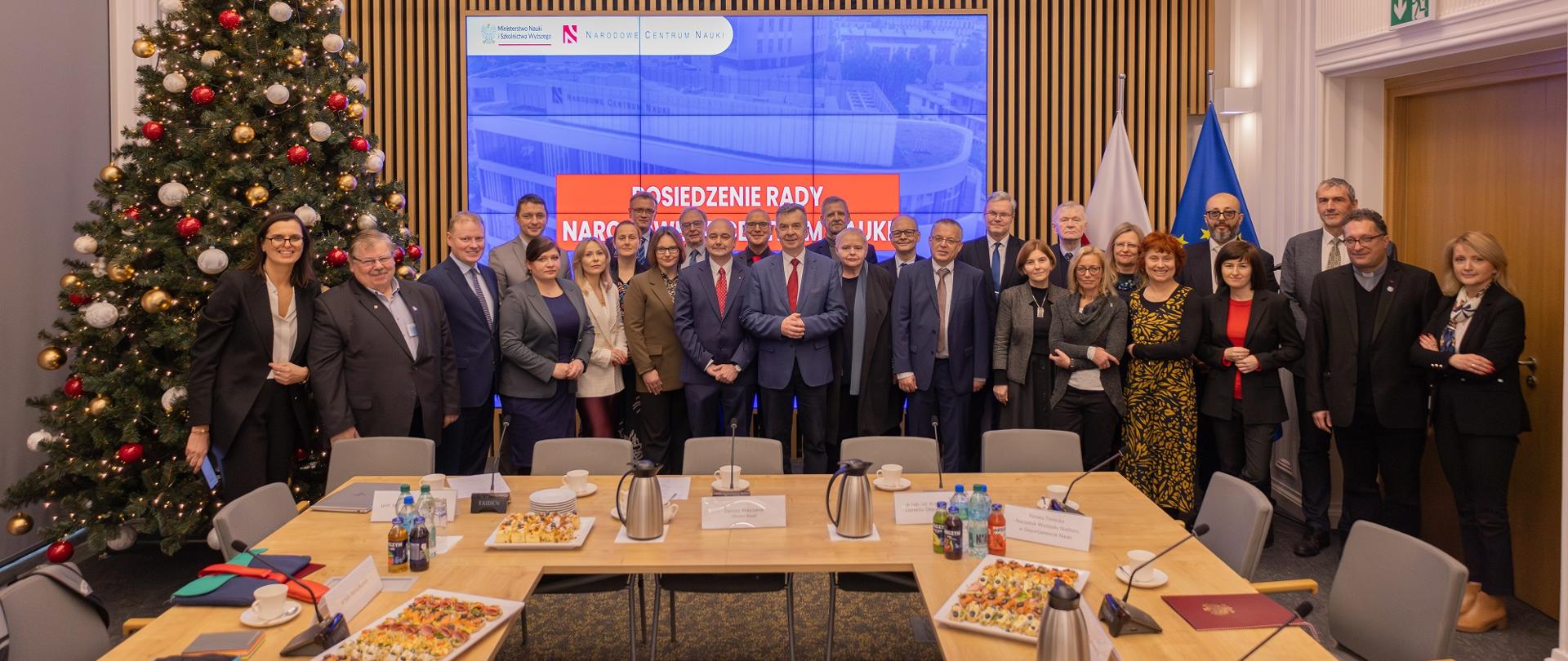 Zdjęcie zbiorowe, przed ścianą z pionowych listewek stoi duża grupa osób, pośrodku minister Wieczorek, za nimi z lewej choinka, pośrodku wbudowany w ścianę wielki ekran, z prawej flagi Polski i UE, przed nimi ustawione w czworokąt stoły.