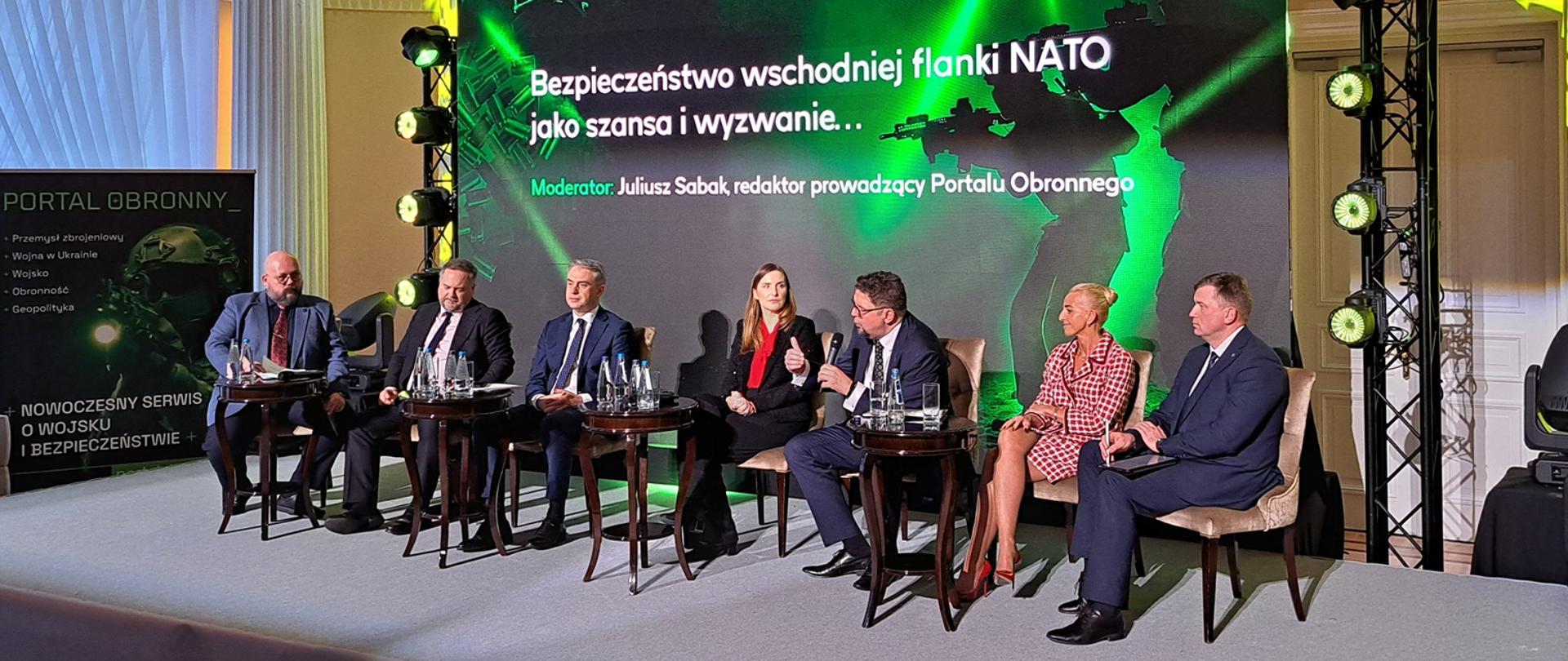 Zdjęcie z boku pod kątem, przed czarno-zieloną ścianką z napisem "Bezpieczeństwo wschodniej flanki NATO jako szansa i wyzwanie" na krzesłach siedzi siedem osób, trzeci od prawej wiceminister Szeptycki mówi do trzymanego w ręku mikrofonu.