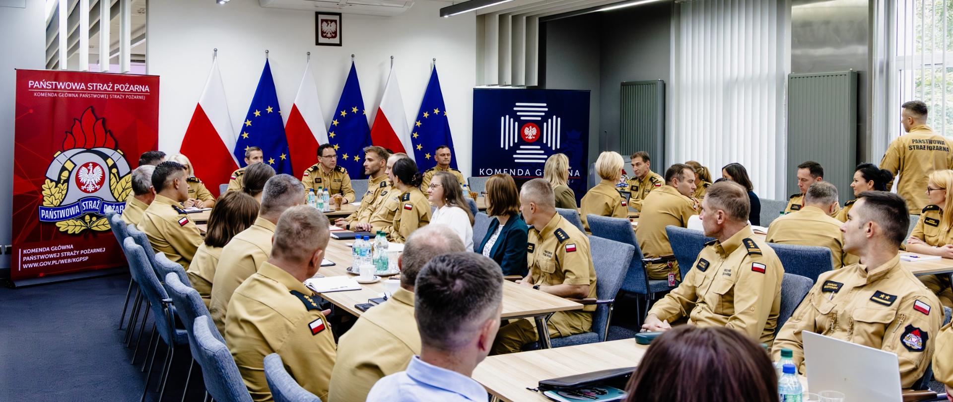 Widok sali konferencyjnej podczas spotkania z udziałem licznej grupy funkcjonariuszy Państwowej Straży Pożarnej. Uczestnicy siedzą przy stołach, a w tle widoczne są flagi Polski i Unii Europejskiej, godło państwowe oraz elementy identyfikacji wizualnej Komendy Głównej PSP.