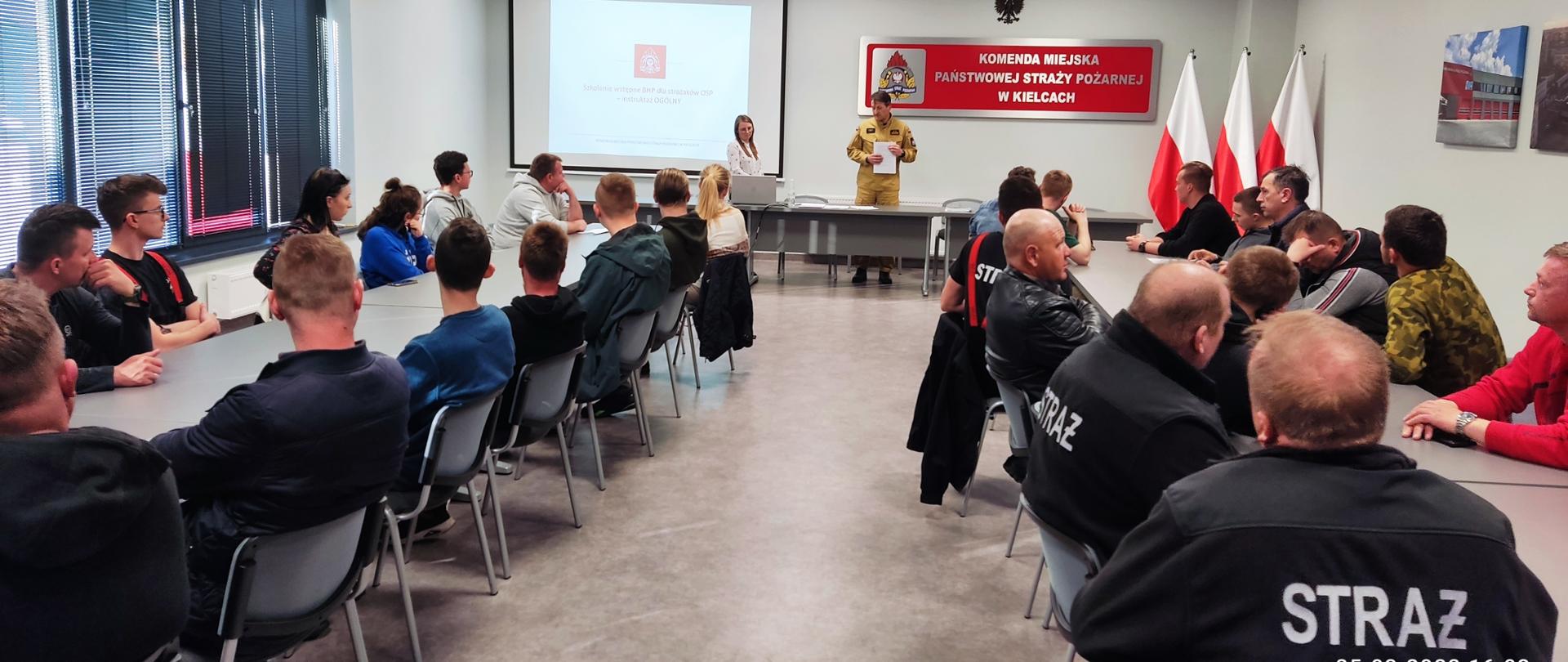 Na zdjęciu przedstawiono: salę wykładową. Przy dwóch rzędach stolików siedzą kursanci, a w szczycie sali witający uczestników zastępca komendanta miejskiego Państwowej Straży Pożarnej w Kielcach bryg. Mariusz Góra i specjalista BHP pani inż. Katarzyna Klepacz.