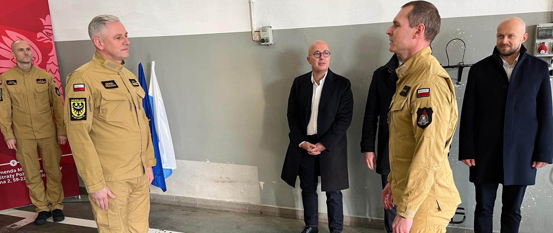 Zdjęcie przedstawia oficjalny meldunek składany podczas uroczystości w hali garażowej jednostki straży pożarnej. Na pierwszym planie dwóch strażaków w piaskowych mundurach służbowych stoi naprzeciw siebie w postawie zasadniczej. Funkcjonariusz po lewej stronie ma na ramieniu naszywkę z dolnośląskim orłem oraz polską flagę, natomiast strażak po prawej stronie melduje gotowość do rozpoczęcia lub zakończenia ceremonii. W tle, przy szarej ścianie, stoi trzech obserwatorów: jeden strażak w piaskowym mundurze oraz dwóch mężczyzn w cywilnych, ciemnych płaszczach, którzy z powagą przyglądają się procedurze. Po lewej stronie widoczny jest fragment czerwonej ścianki z logo Państwowej Straży Pożarnej oraz biało-niebieska flaga. Całość odbywa się na betonowej posadzce z namalowaną białą linią, co podkreśla formalny i zdyscyplinowany charakter wydarzenia.