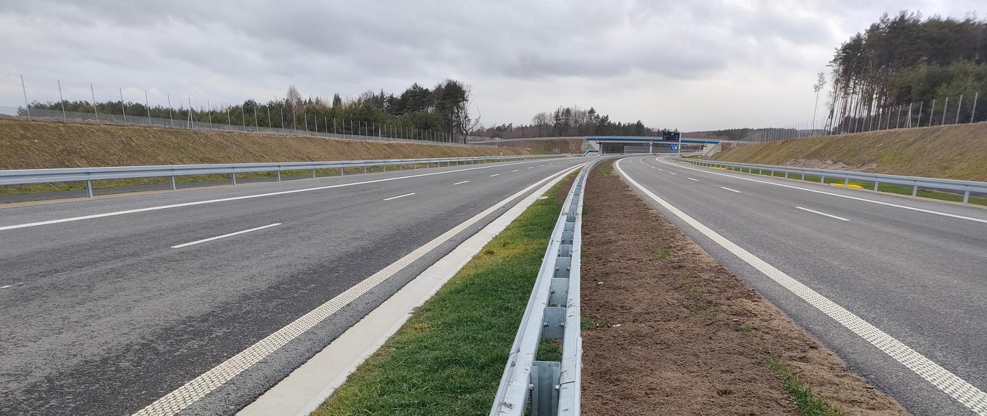 Widok na drogę dwupasmową rozdzieloną pasem zieleni i barierami energochłonnymi. W tle widać wiadukt nad drogą. 