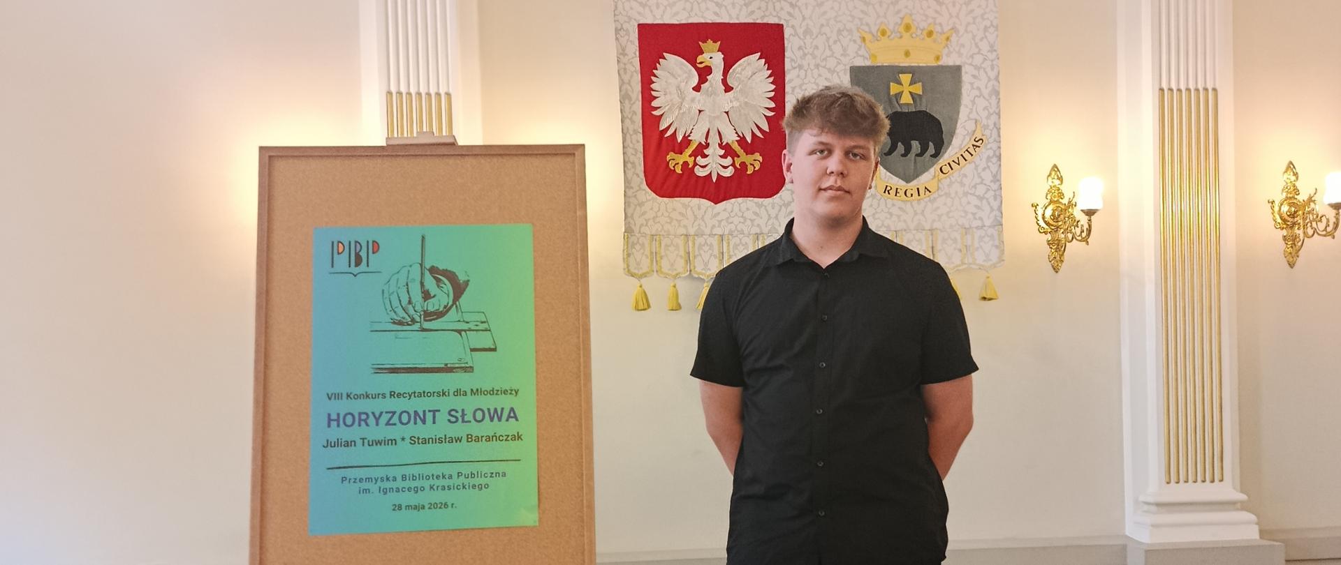 Zdjęcie przedstawia aulę biblioteki, na scenie stoi uczeń ubrany na czarno, obok na sztaludze zawieszony jest zielony plakat z napisem VIII Konkurs Recytatorski „Horyzont Słowa”, w tle biała ściana z godłem i herbem Przemyśla.