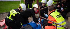Widok na konkurencję. Strażacy zajmujący się osobą poszkodowaną leżącą na ziemi – asekurując ją i opatrując rany. W tle organizator i osoby oceniające poprawność przebiegu konkurencji.