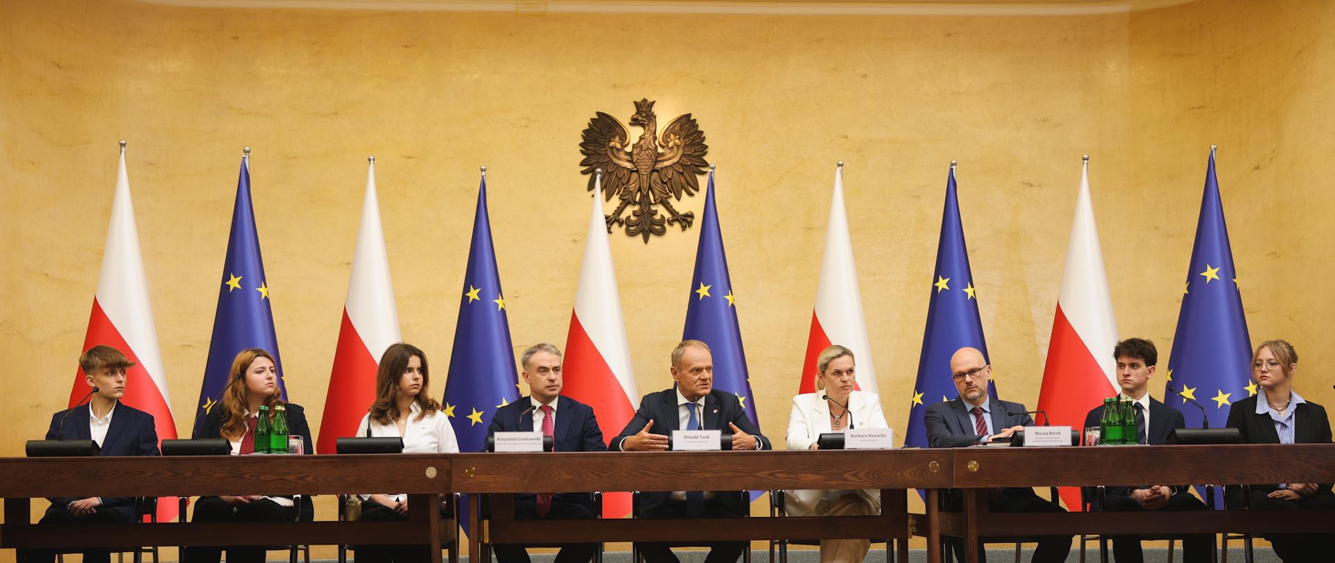 9 osób, w tym Premier Donald Tusk i ministrowie, siedzą przy brązowym stole. Przed nimi stoją mikrofony, butelki z wodą oraz szklanki. Za nimi stoją biało-czerwone i unijne flagi, nad którymi góruje godło Polski.
