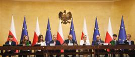 9 osób, w tym Premier Donald Tusk i ministrowie, siedzą przy brązowym stole. Przed nimi stoją mikrofony, butelki z wodą oraz szklanki. Za nimi stoją biało-czerwone i unijne flagi, nad którymi góruje godło Polski.