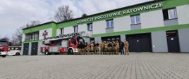 Pamiątkowe zdjęcie grupy kilkunastu mężczyzn w mundurach strażackich i ratowniczych przed nowoczesną siedzibą Górniczego Pogotowia Ratowniczego. Ratownicy stoją w rzędzie na wybrukowanym placu obok specjalistycznego wozu strażackiego z rozłożoną częściowo drabiną. Budynek jest szaro-zielony, z widocznymi bramami garażowymi i dużym napisem na elewacji.