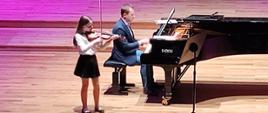 Dziewczynka gra na skrzypcach na scenie sali koncertowej stojąc po lewej stronie przed fortepianem, na którym gra mężczyzna. Z przodu widać od tyłu publiczność siedzącą na widowni.