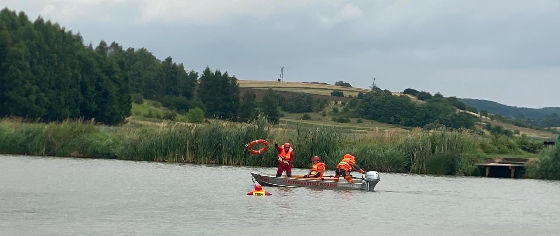 Na zdjęciu trzech ratowników na łodzi jeden rzuca kołem ratunkowej s w stronę osoby topiącej się