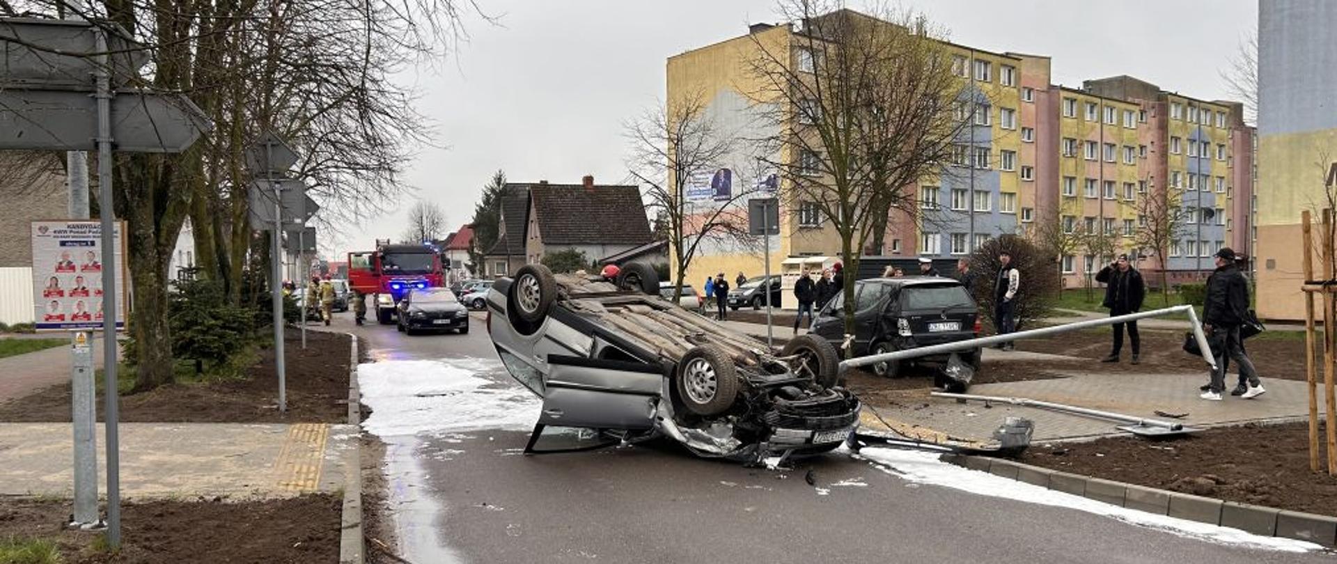 Widok na ulicę. W tle bloki i gapie. Na pierwszym planie srebrne auto na dachu. Pod autem poduszka piany. Zdjęcie wykonane w dzień.