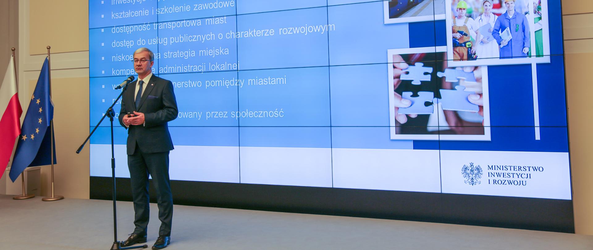 Podczas konferencji prasowej minister Jerzy Kwieciński stoi na scenie przed mikrofonem. Za jego plecami na dużym ekranie wyświetlona jest prezentacja. W lewym rogu stoją dwie flagi Polski i UE.