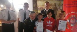 Zastępca Opolskiego Komendanta Wojewódzkiego PSP w Opolu st. bryg. Wojciech Nawara, Wiceprezes Zarządu Głównego Związku Ochotniczych Straży Pożarnych Rzeczypospolitej Polskiej dh Teresa Tiszbierek, Prezes Zarządu Oddziału Wojewódzkiego Związku Ochotniczych Straży Pożarnych Rzeczypospolitej Polskiej Województwa Opolskiego dh Andrzej Borowski oraz Sekretarz Zarządu Oddziału Wojewódzkiego Związku Ochotniczych Straży Pożarnych Rzeczypospolitej Polskiej Województwa Opolskiego dh Norbert Waliczek, stoją do wspólnego zdjęcia z finalistami etapu wojewódzkiego Ogólnopolskiego Turnieju Wiedzy Pożarniczej "Młodzież zapobiega pożarom" w I grupie wiekowej.